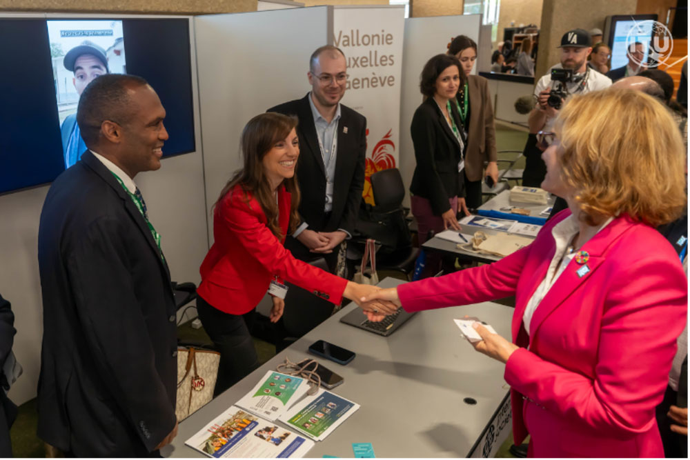 two women shake hands at a booth, standing with a few other people.