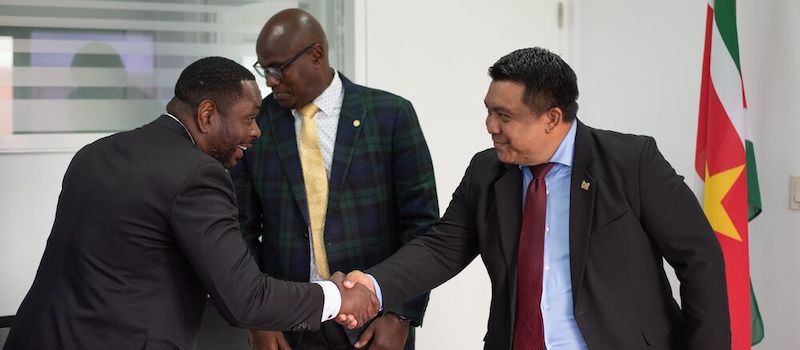 Dos hombres dándose la mano sonriendo con la bandera de Surinam en segundo plano.