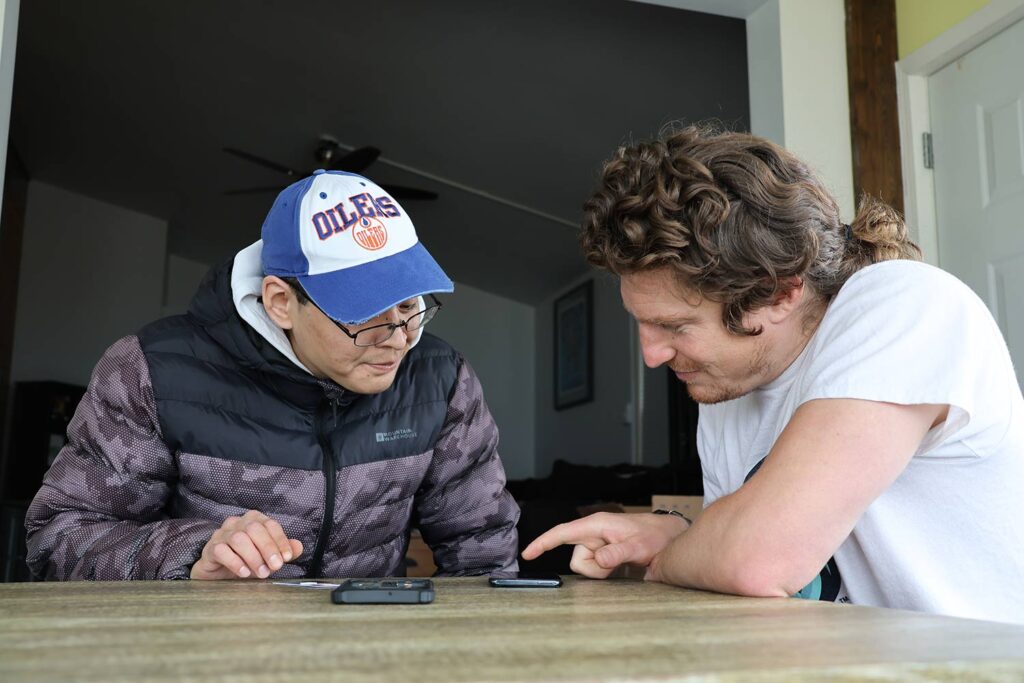 Two people sit at a table as one shows the other how to set up a new SIM card on their phone.