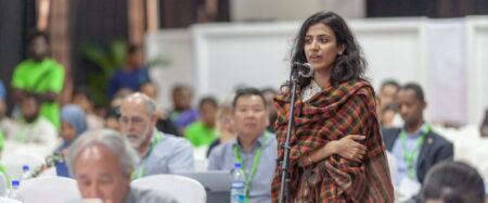 Woman in green and black scarf giving a speech at a conference.