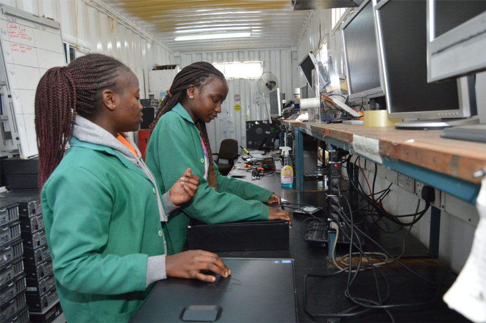 Two women in green jackets stand in front of computers